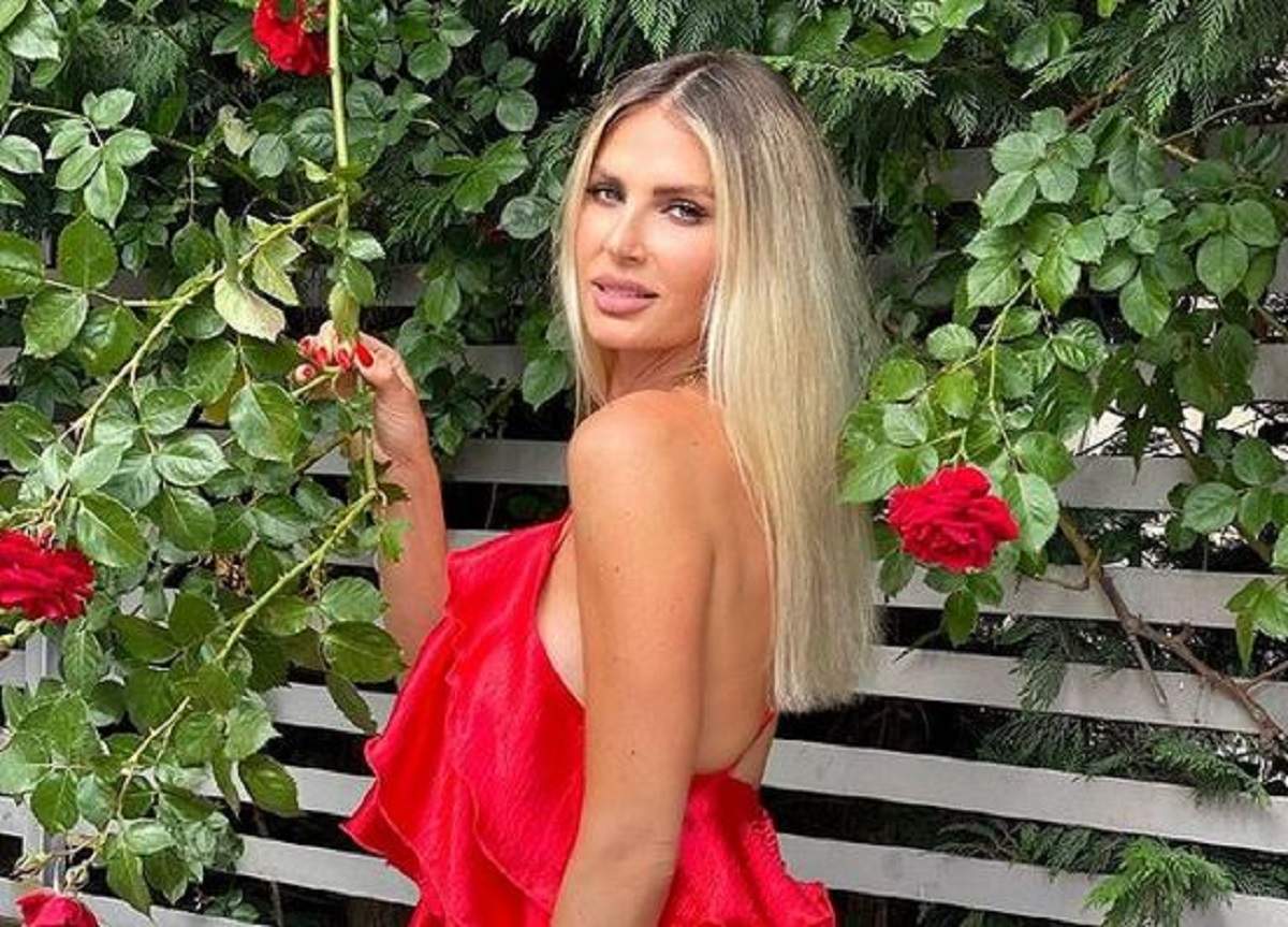 Andreea Bănică poartă o rochie roșie și zâmbește. Vedeta ține în mână tulpina unui trandafir.