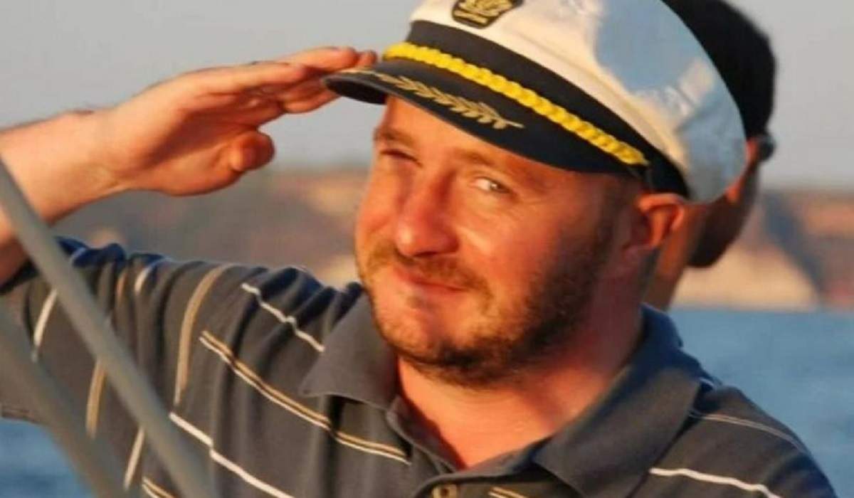 Fostul jurnalist Alin Bogdan poartă tricou gri cu dungi albe și galbene și o șapcă albă, de marinar. Acesta salută cu mâna la nivelul capului.