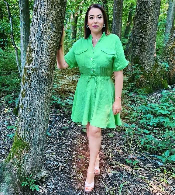 Oana Roman se ține cu o mână de un copac, în parc. Vedeta poartă rochie verde și sandale albe.