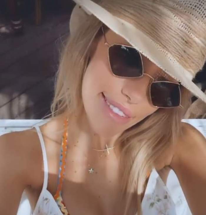 Gabriela Prisăcariu își face un selfie de la mare, poartă ochelari de soare negri, pălărie de paie și rochie albă,