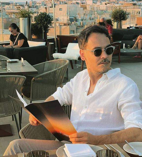 Răzvan Simion stă la terasă în Grecia, are meniul în mână și poartă ochelari de soare și cămașă albă.