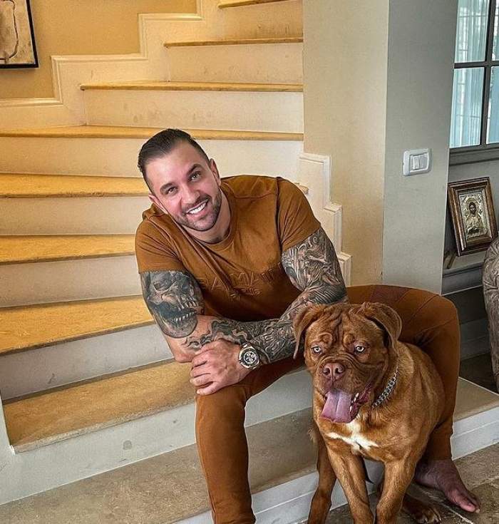 Alex Bodi stă pe scări, zâmbește și poartă tricou și pantaloni maro. Lângă el e câinele lui.