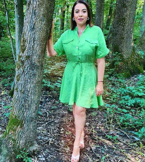 Oana Roman se ține cu o mână de un copac, în parc. Vedeta poartă rochie verde și sandale albe.