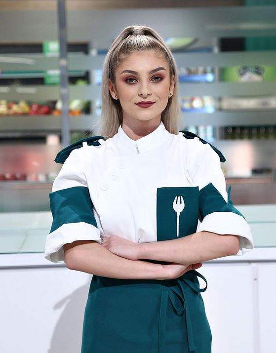 Elena Matei poartă uniformă de bucătar albă cu verde și e în bucătăria Chefi la cuțite. Fosta concurentă zâmbește și are mâinile încrucișate.