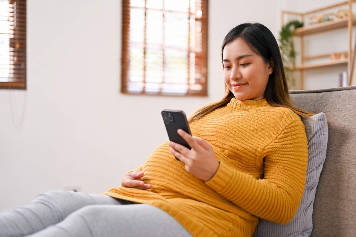 Vedere laterală a unei femei gravide asiatice fericite care citește blogul despre maternitate pe internet, verifică data nașterii copilului ei, comandă lucruri pentru bebeluș la cumpărături online