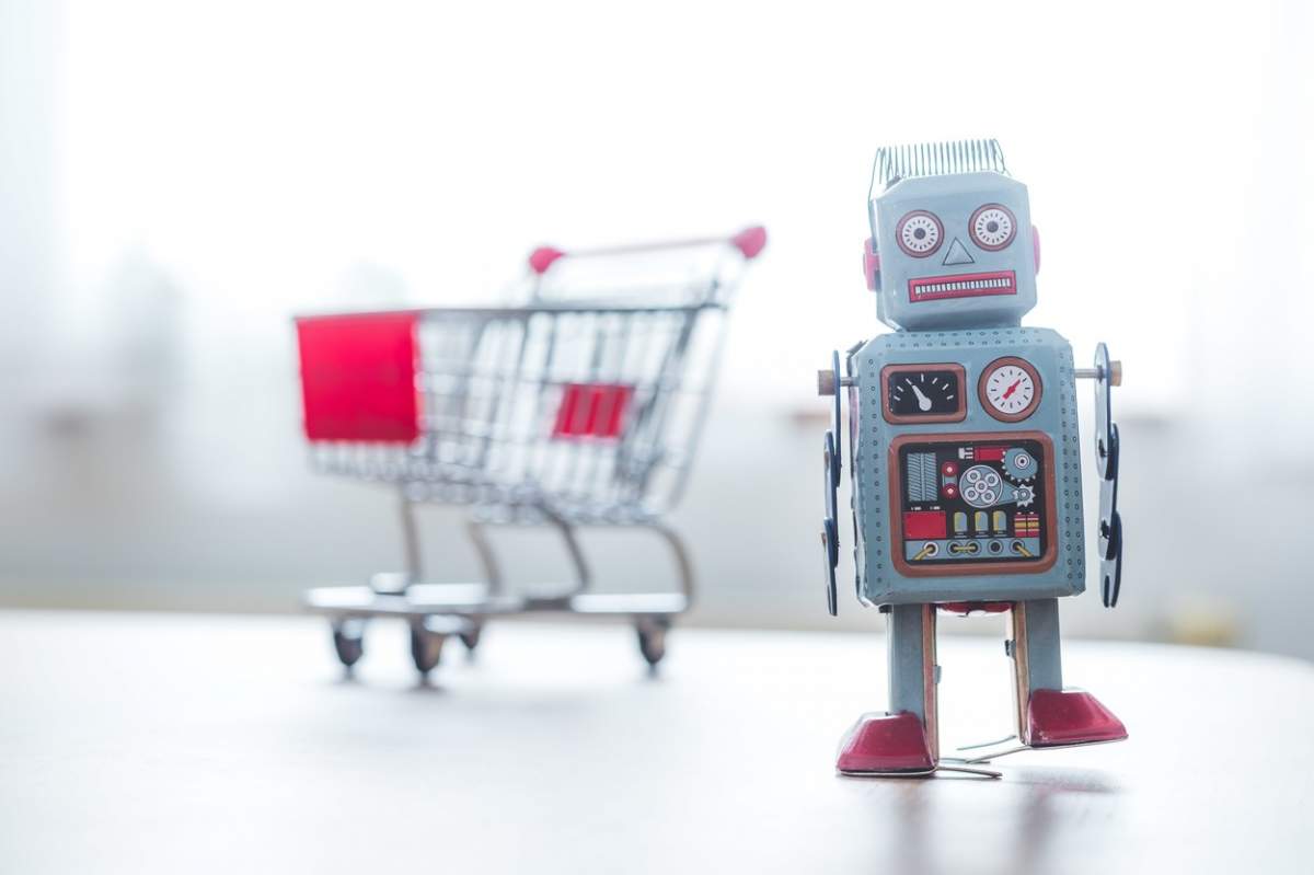 Conceptul de algoritm de cumpărături online. Robot jucărie pe birou, coș de cumpărături în fundal neclar