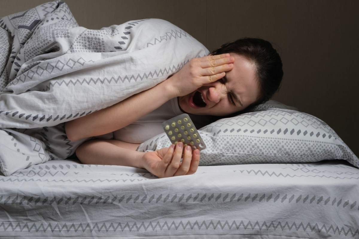 O femeie cu insomnie stă întinsă în pat acoperită cu o pătură, căscă și ia pastile pentru un somn sănătos