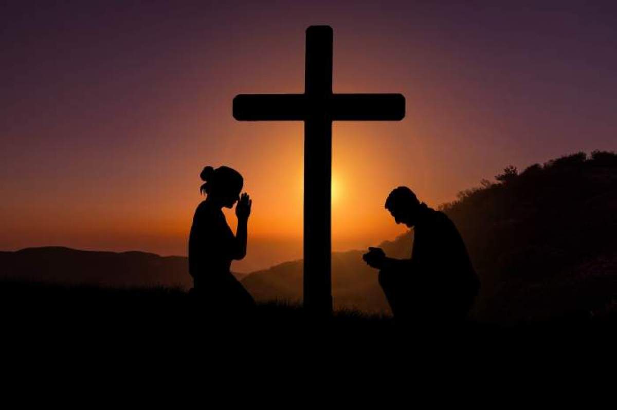 o cruce și doi oameni care se roagă lângă ea.