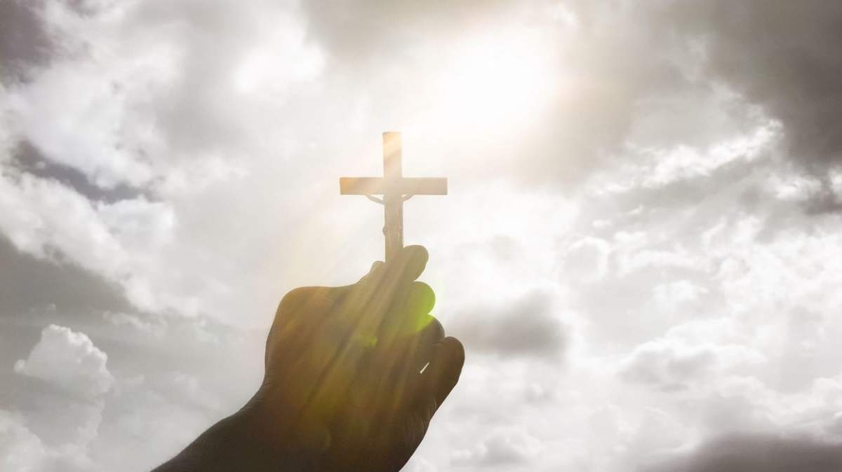 mână ținând Sfânta Cruce cu fundal strălucitor de cer. Conceptul de creștinism.