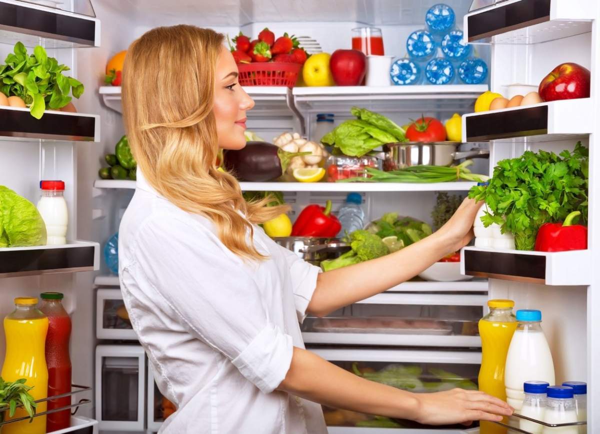 Fotografie cu o femeie care stă lângă frigider și frigiderul plin cu fructe și legume