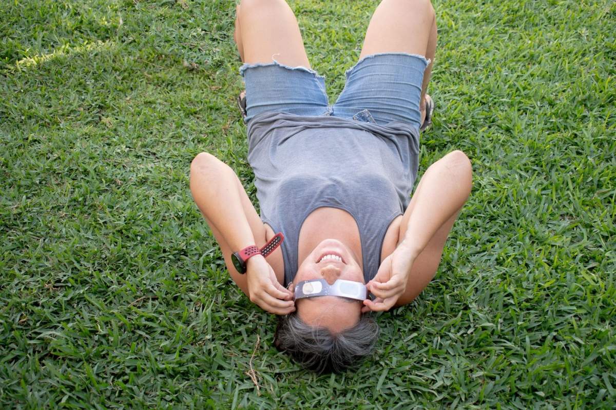 O persoană întinsă pe iarbă privind cerul, folosind ochelari cu filtre solare pentru a vedea eclipsa