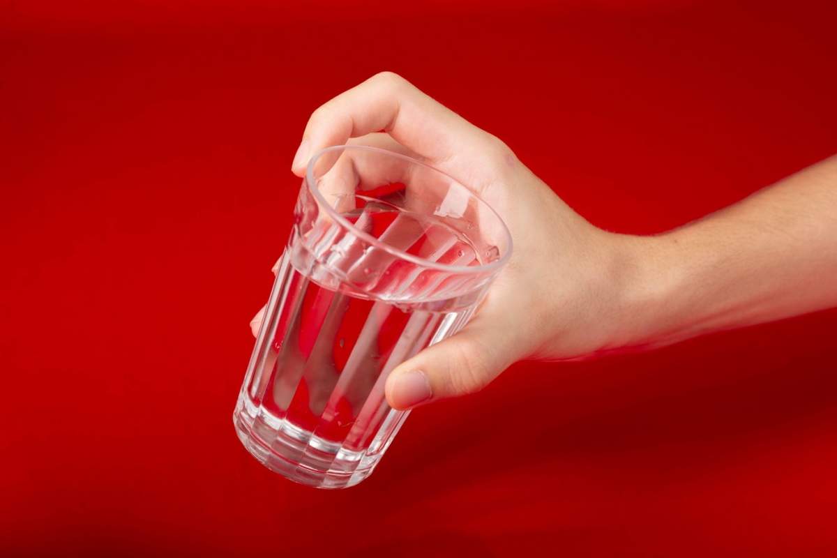 Mână de bărbat care ține un pahar de sticlă cu apă înăuntru. Izolat pe fundal roșu