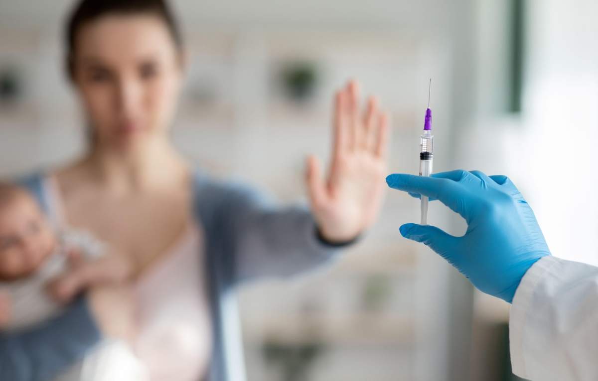 Ezitare la vaccinare. Femeie cu nou-născut care refuză seringa în mâna medicului.