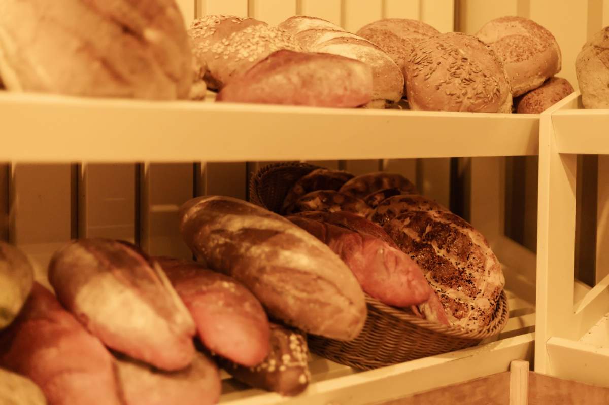 O varietate de pâini artizanale sunt aranjate cu grijă pe rafturi de lemn într-o brutărie confortabilă