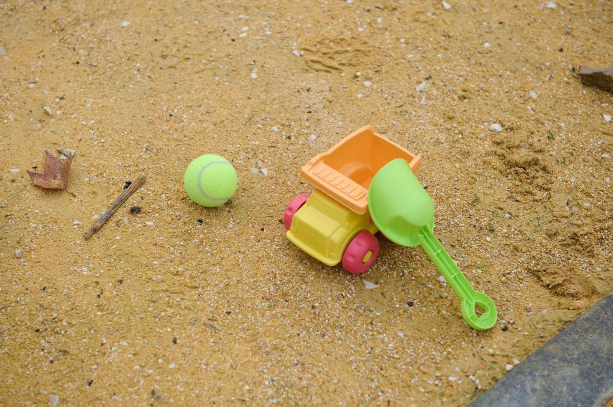 Scenă cu nisip strălucitor, cu o lopată verde, o găleată portocalie, un camion galben și o minge de tenis. Ideală pentru familie, joacă în aer liber, distracție de vară și activități pentru copii.