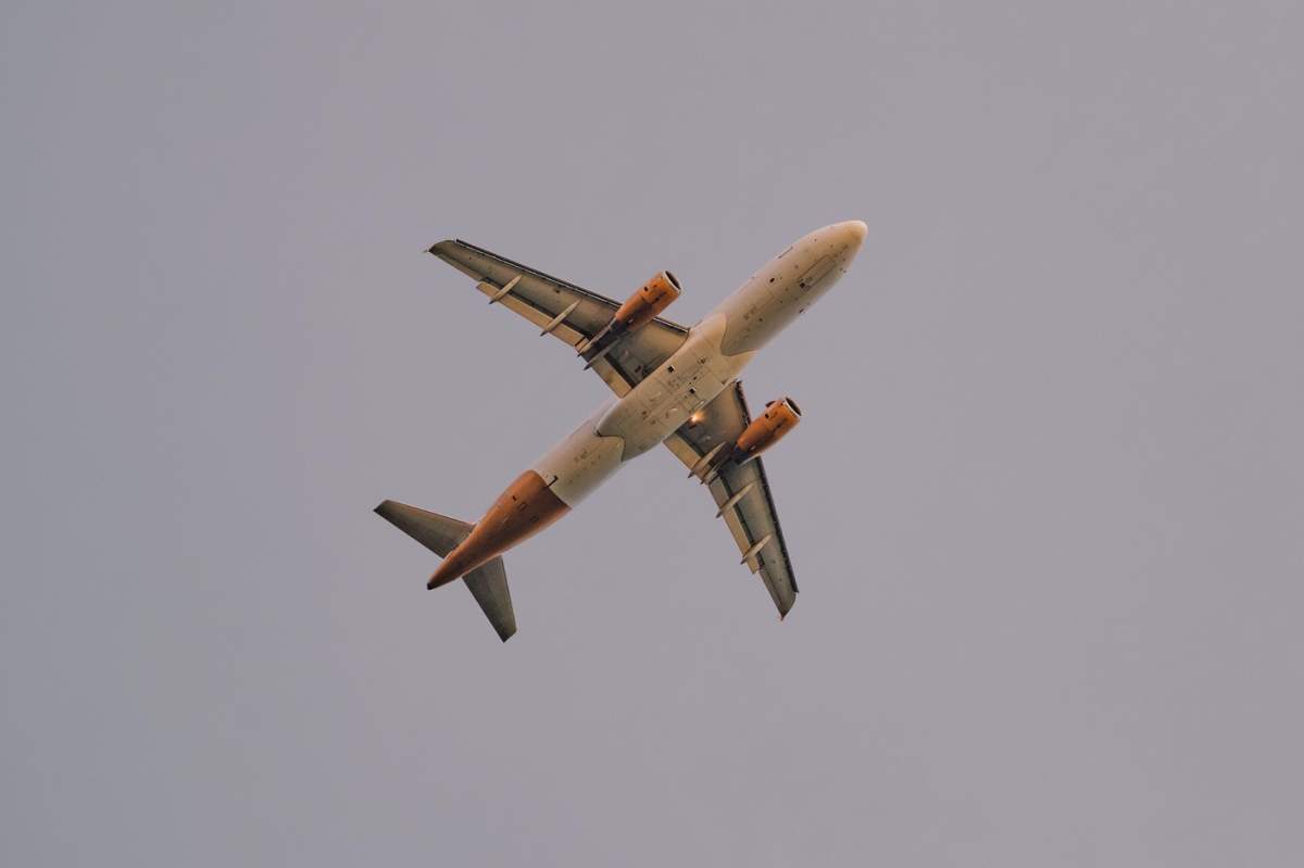 Avion comercial văzut direct de jos, arătând fuselajul, motoarele și numărul de cod pe un cer înnorat.