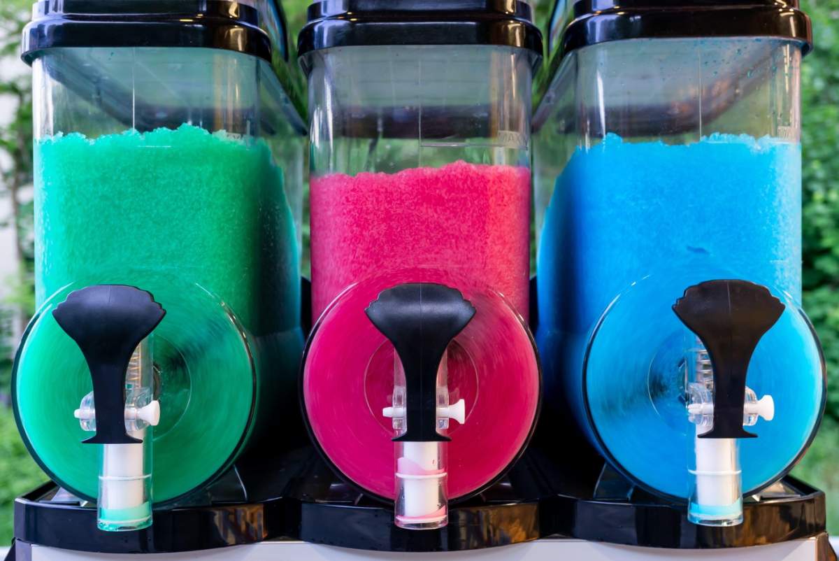 Trei recipiente colorate pentru băuturi înghețate, umplute cu gheață măcinată în culori verde, roz și albastru, gata pentru a fi servite într-o zi toridă.