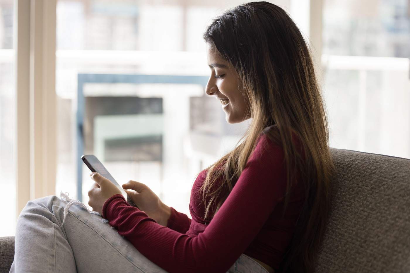 O femeie indiană zâmbitoare își ia o pauză așezată pe o canapea cu un smartphone modern, navigând prin aplicații fără țintă, petrece timpul liber pe internet, derulează fluxul de știri, descarcă o nouă aplicație mobilă amuzantă acasă