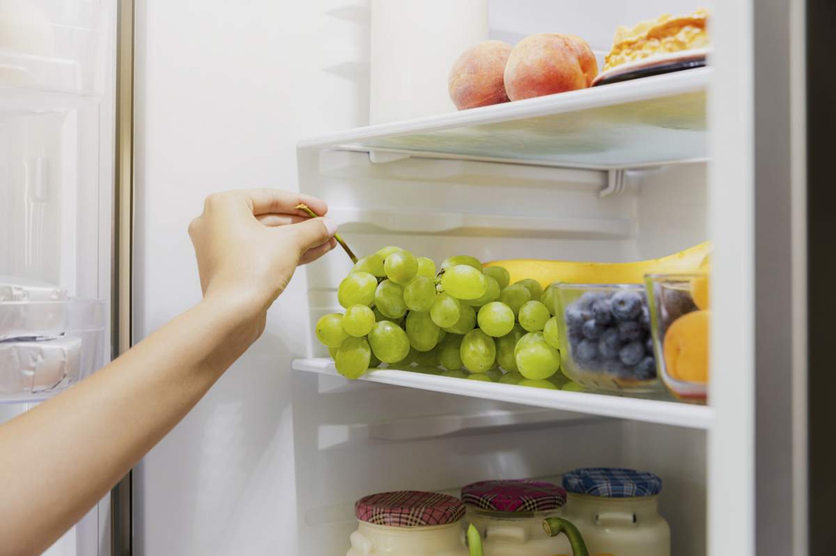 Femeie care ia, apucă sau ridică un ciorchine de struguri verzi de pe raftul deschis al frigiderului sau sertarul frigiderului plin cu fructe, afine, sticle cu iaurt. Dietă sănătoasă, concept de stil de viață