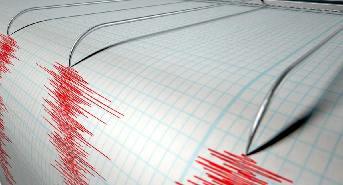 Prim-planul acului unui seismograf care trasează o linie roșie pe hârtie milimetrică, reprezentând activitatea seismică și cutremurelor pe o izolație