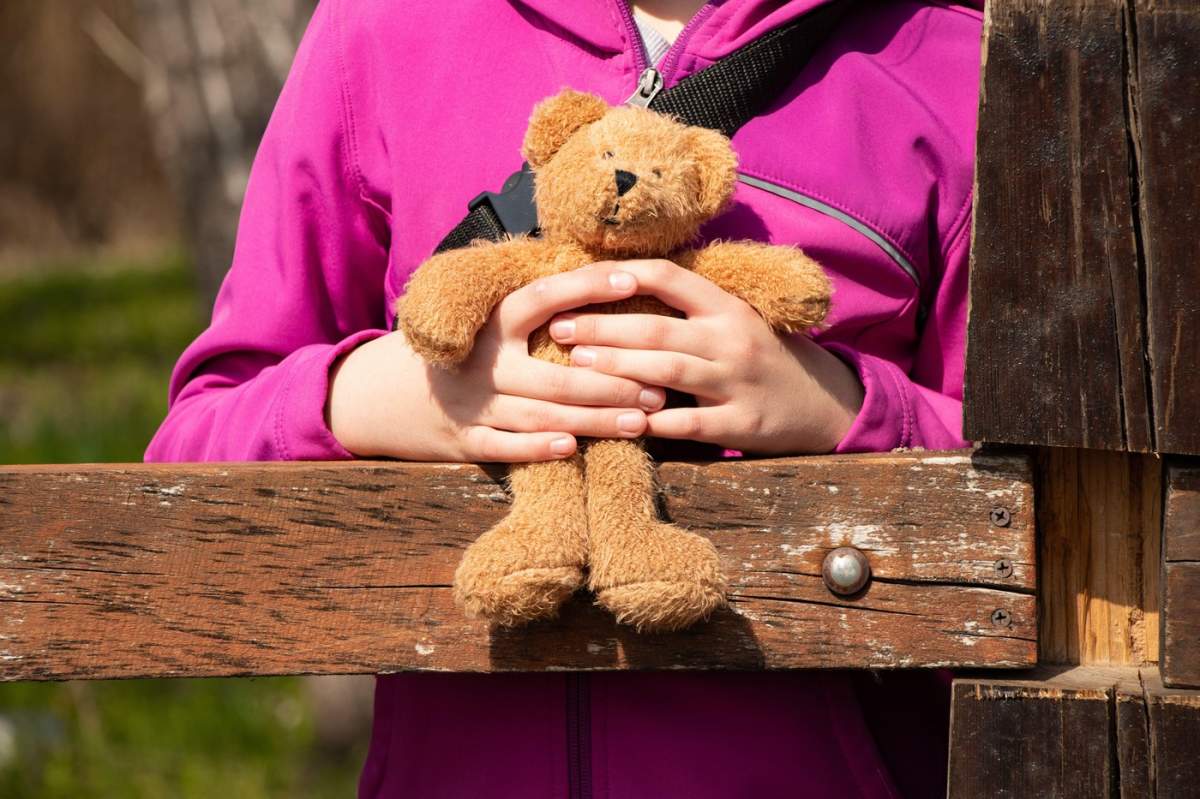 Fată care ține un ursuleț de pluș așezată pe un gard primăvara într-un parc din Ucraina, la soare
