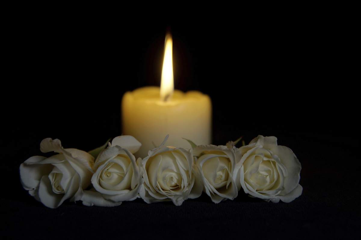 Trandafiri albi frumoși cu o lumânare aprinsă pe fundal întunecat. Floare funerară și lumânare pe masă pe fundal negru, spațiu pentru copie. Simbol funerar Covid-19. Concept de card de condoleanțe și dispoziție.