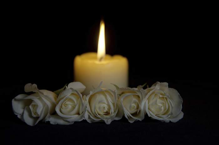 Trandafiri albi frumoși cu o lumânare aprinsă pe fundal întunecat. Floare funerară și lumânare pe masă pe fundal negru, spațiu pentru copie. Simbol funerar Covid-19. Concept de card de condoleanțe și dispoziție.