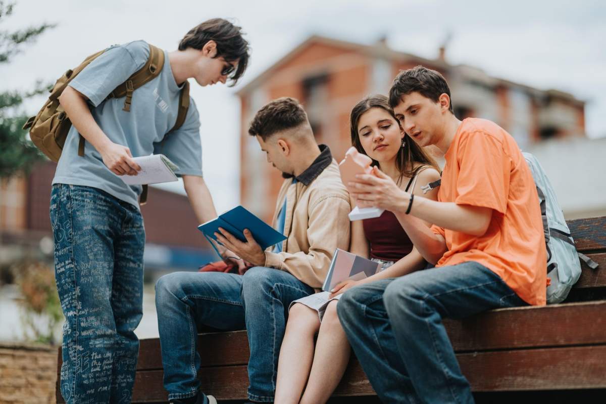 Patru studenți care studiază împreună în aer liber pe o bancă din campusul universitar cu cărți și telefon
