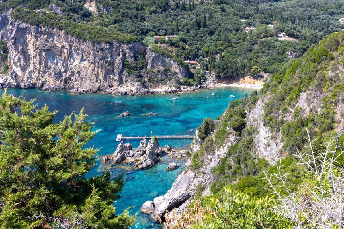 Golf de pe insula Corfu, cu ape turcoaz cristaline, plajă cu nisip, stânci și un dig, sub cerul însorit de vară al Greciei.