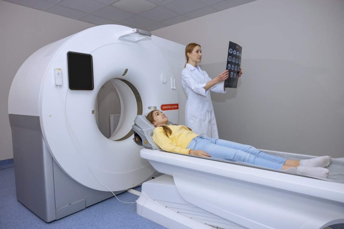 O tânără doctoriță blondă examinează o imagine RMN într-o clinică, stând lângă aparatul RMN și o pacientă micuță și drăguță.
