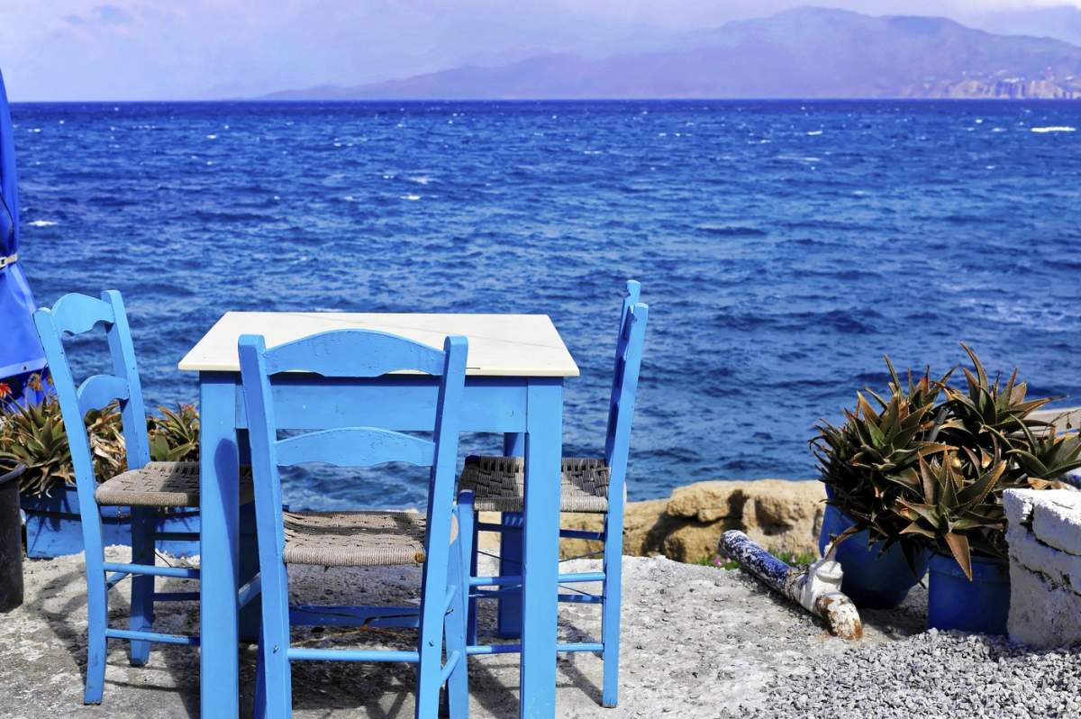 Tavernă, golf, plaja Matala, Matala, Creta, Grecia; masă și scaune albastre cu vedere spre marea azurie și coastă în fundal, Creta, Grecia.
