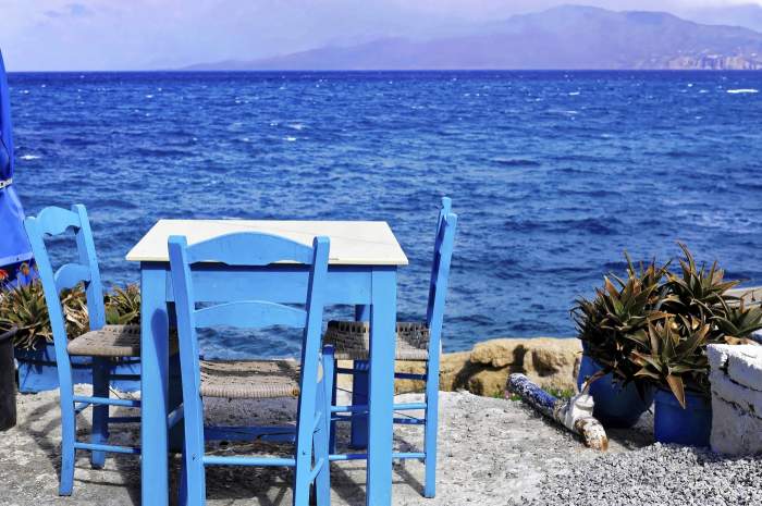 Tavernă, golf, plaja Matala, Matala, Creta, Grecia; masă și scaune albastre cu vedere spre marea azurie și coastă în fundal, Creta, Grecia.