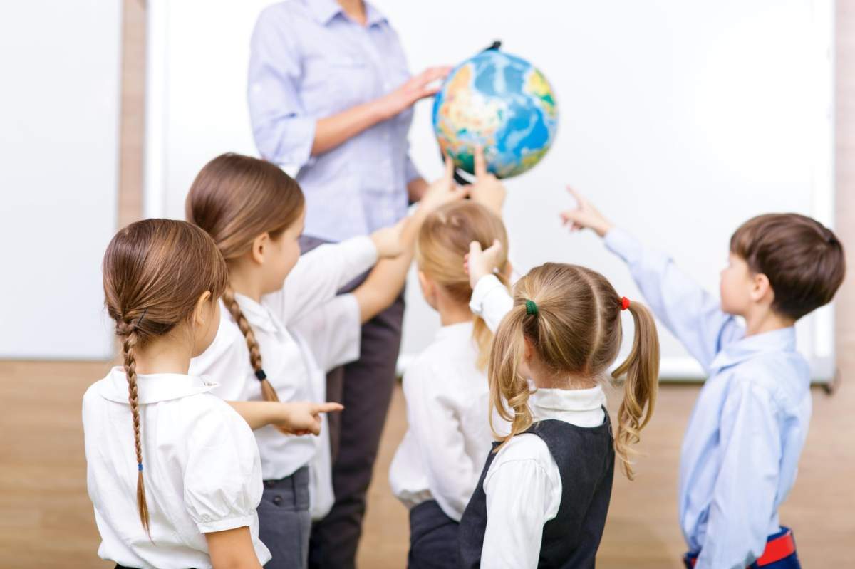 Exercițiu de geografie. Un grup de elevi mici pare entuziasmat și arată cu degetul spre globul pământesc pe care îl ține profesorul lor.