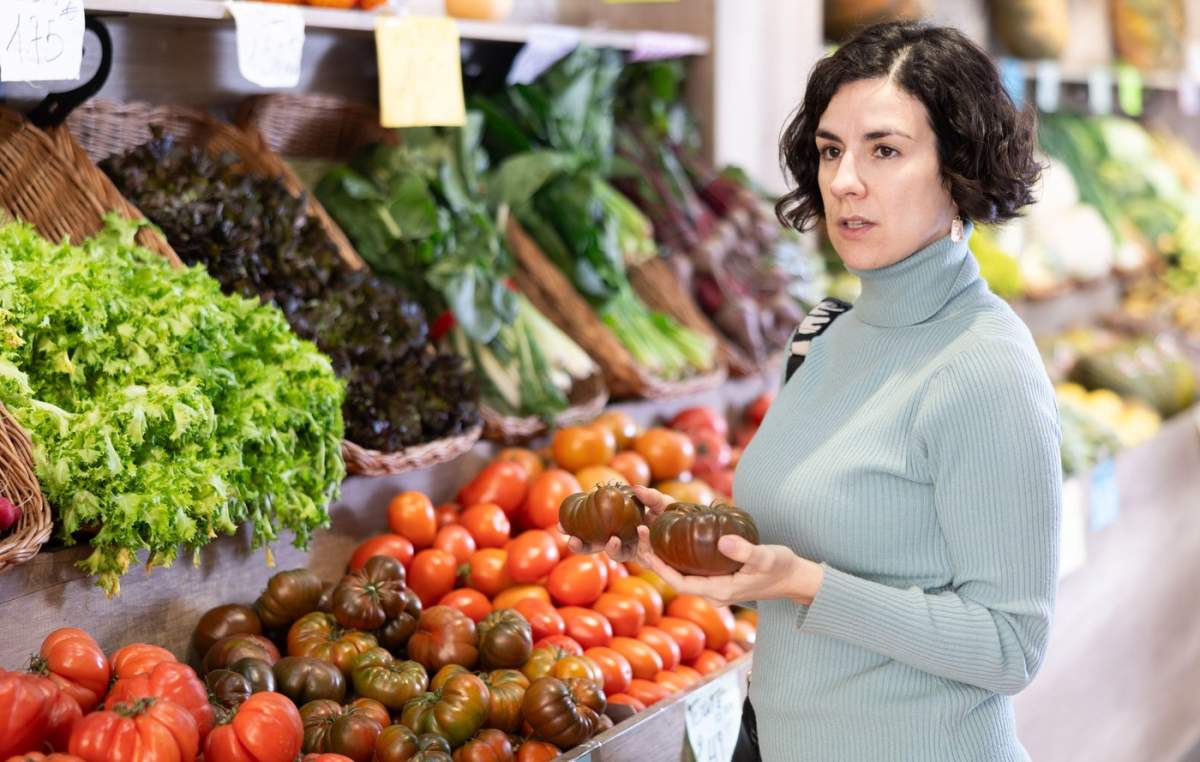 O femeie de vârstă mijlocie cumpără roșii roșii și coapte dintr-un supermarket. O femeie europeană stă lângă raionul de legume, ține roșiile în mâini, le examinează și le alege pentru a găti cina.