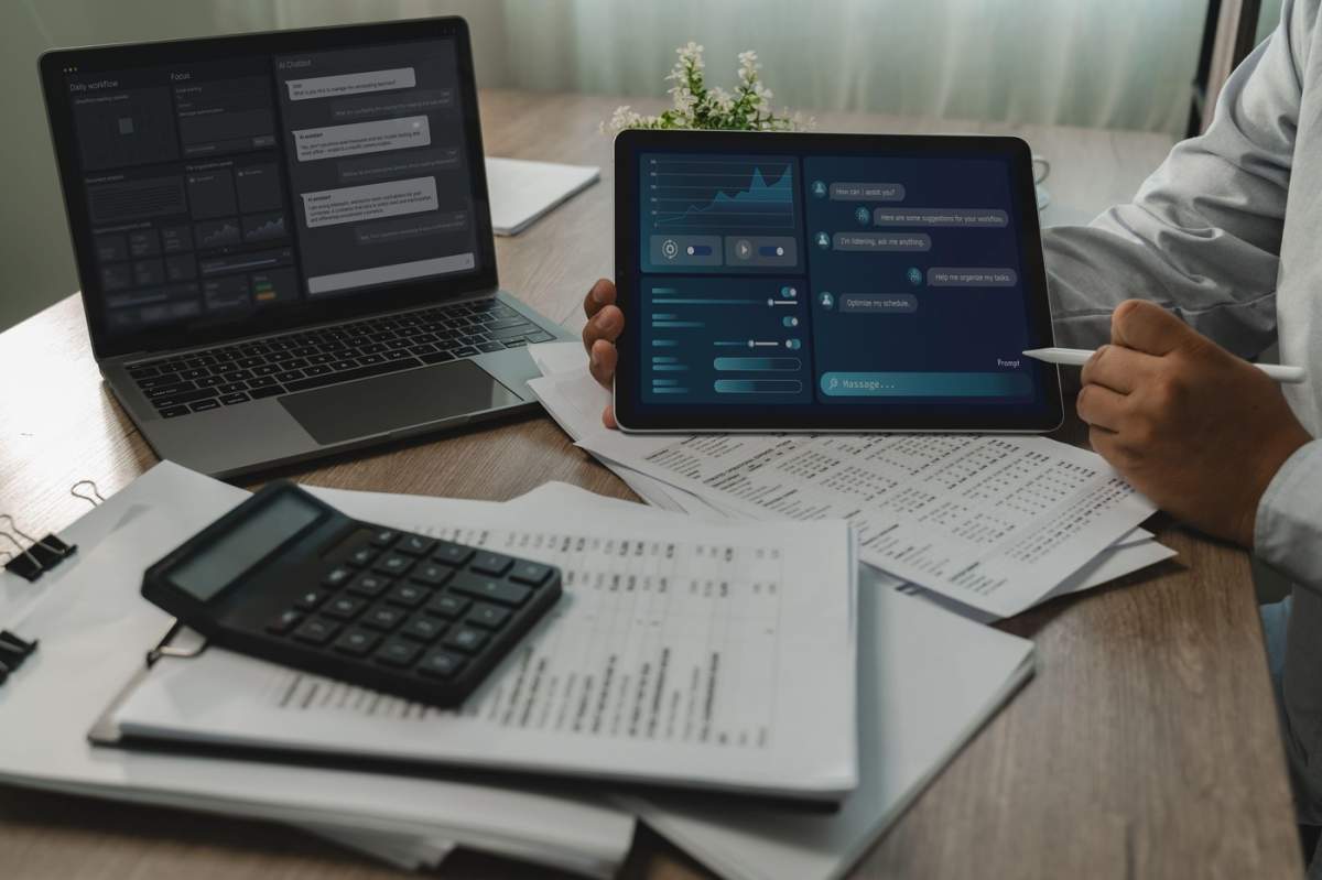 O persoană folosește interfețe de asistență AI pe un laptop și o tabletă deasupra unor hârtii și a unui calculator, organizând sarcinile cu un stylus și susținând un flux de lucru digital de acasă la birou.