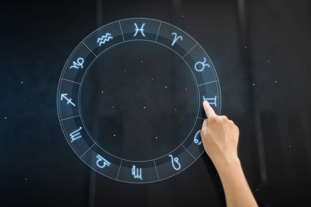Concept legat de tehnologie, astrologie și horoscop – o mână utilizează un panou interactiv cu semnele zodiacale, având ca fundal cerul nocturn și stelele.